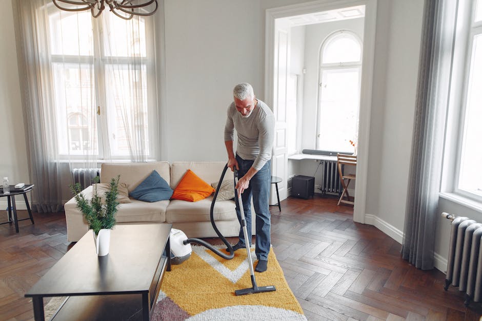 A young boy is vacuuming a beige fabric sofa in a living room with wooden flooring and a mustard-colored rug, while an older man with gray hair and beard is lying on the sofa, appearing relaxed with h