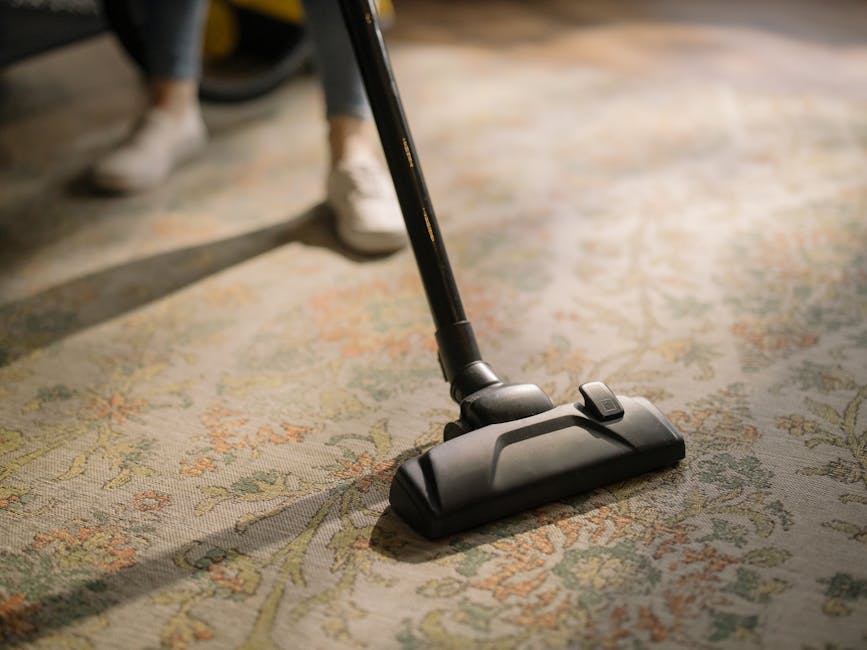 A close-up image of a black carpet vacuum cleaner head positioned on an ornate, patterned carpet with floral motifs in muted colors. The vacuum hose extends upward, with a person's legs visible in the
