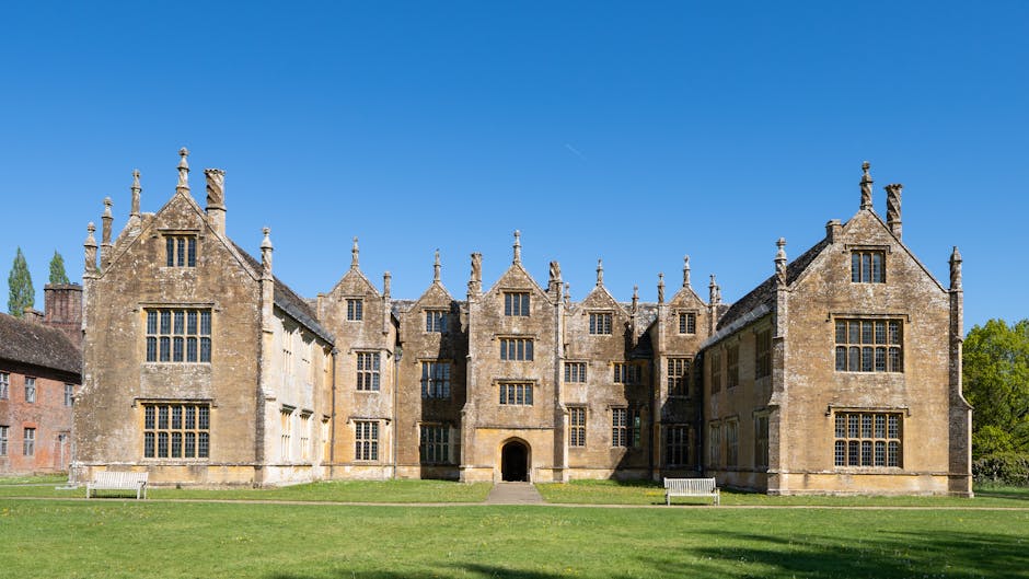 A large historic mansion with a yellow facade and multiple turrets, surrounded by lush green trees and a well-maintained lawn, captured in bright daylight. The building features numerous windows and a